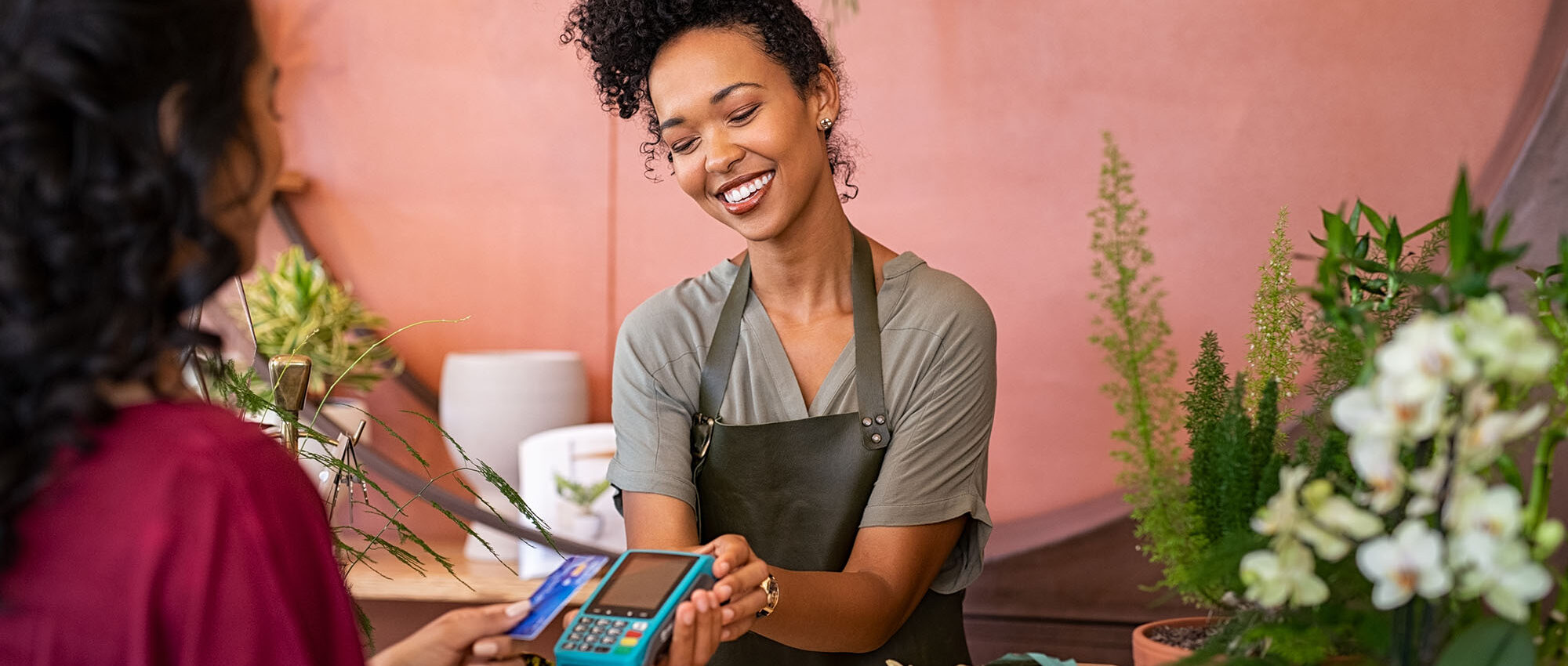 Smiling florist holding card reader machine at counter with customer paying with credit card. Young african american florist shop assistant holding payment machine while buyer purchase a bunch flower.