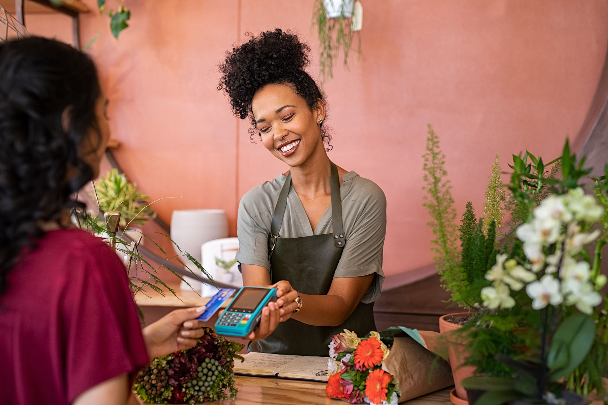 Smiling florist holding card reader machine at counter with customer paying with credit card. Young african american florist shop assistant holding payment machine while buyer purchase a bunch flower.